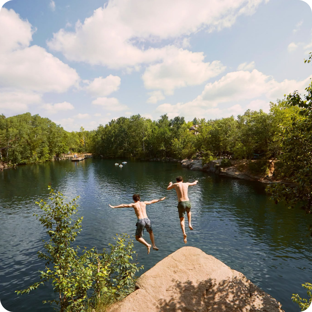 Two young men jump off a rock into a clear lake surrounded by trees under a partly cloudy sky.
